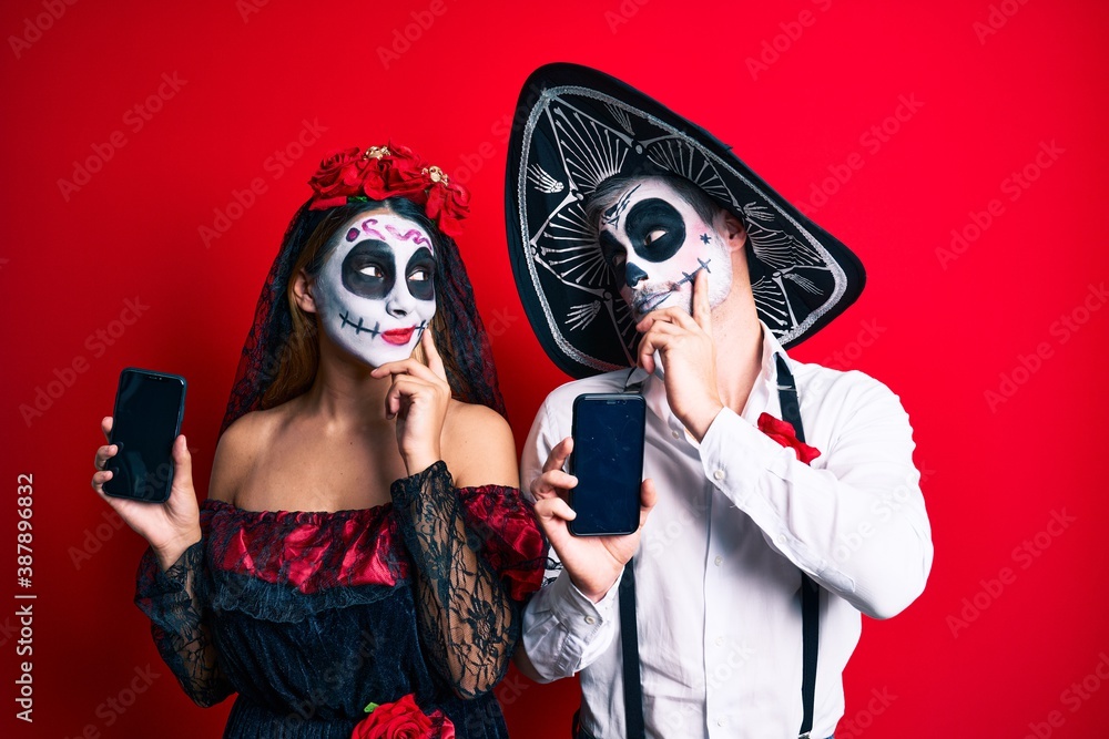 Couple wearing day of the dead costume holding smartphone showing screen serious face thinking about question with hand on chin, thoughtful about confusing idea Couple wearing day of the dead costume holding smartphone showing screen serious face thinking about question with hand on chin, thoughtful about confusing idea