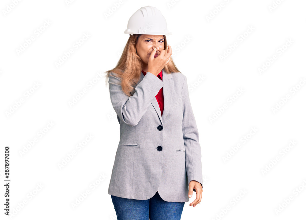 Beautiful young woman wearing architect hardhat smelling something stinky and disgusting, intolerable smell, holding breath with fingers on nose. bad smell