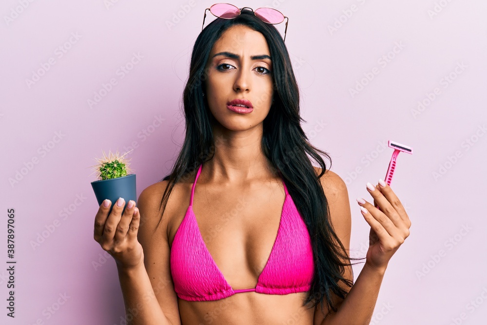 Beautiful hispanic woman wearing bikini holding cactus and razor clueless and confused expression. doubt concept. Beautiful hispanic woman wearing bikini holding cactus and razor clueless and confused expression. doubt concept.