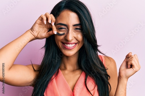 Beautiful hispanic woman holding fake lashes screaming proud, celebrating victory and success very excited with raised arm