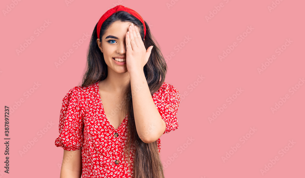 Brunette teenager girl wearing summer dress covering one eye with hand, confident smile on face and surprise emotion. Brunette teenager girl wearing summer dress covering one eye with hand, confident smile on face and surprise emotion.