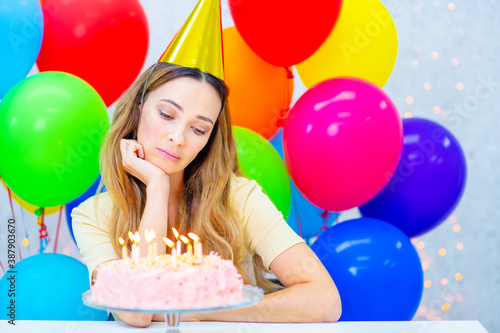 A middle-aged woman in a party cap in front of a birthday cake with candles looks sadly at the candles.