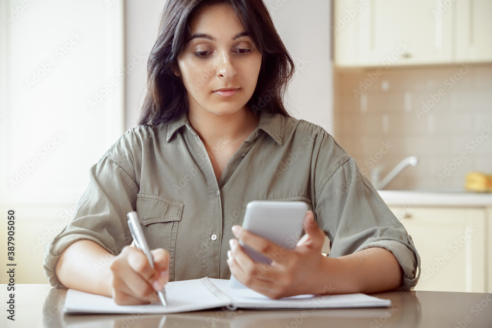 Indian female student holding phone using remote online education mobile app studying at home using smartphone for online learning, watching webinar, video conference calling sitting at desk at home. Indian female student holding phone using remote online education mobile app studying at home using smartphone for online learning, watching webinar, video conference calling sitting at desk at home.