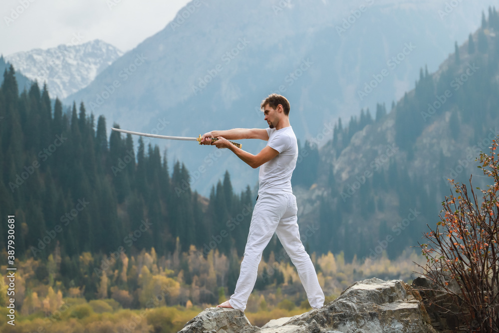 Naklejka premium A young man stands on a large stone and holds a Japanese sword in his hands.