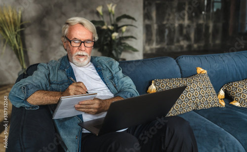 Focused elderly man writing notes in notebook watching webinar or online training using laptop computer, modern senior male with gray hair and beard learning online