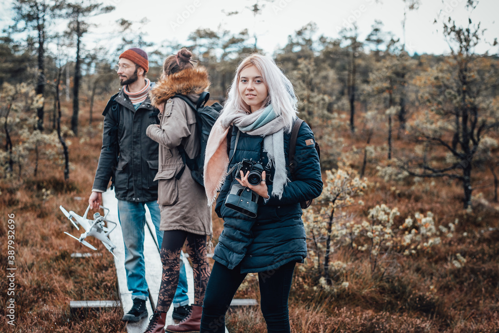 Attractive blond girl with camera poses at wooden path looking at camera behind her friends in lovely and relaxed autumn forest.