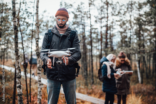 Bearded guy stays holding quadcopter and preparing it behind his friends in autumn lovely forest.
