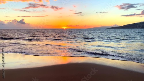 Medium shot of gentle waves rolling to shore in a tropical island sunset with sandy foreground and a distant island in the distance with rich orange and yellow sky horizon.