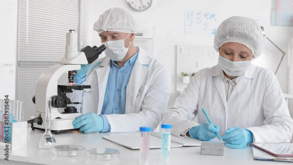 Medium shot of male Caucasian doctor looking through microscope talking to senior Asian female assistant taking notes in journal. Colleagues wearing medical masks, caps, gloves sitting by desk in lab Medium shot of male Caucasian doctor looking through microscope talking to senior Asian female assistant taking notes in journal. Colleagues wearing medical masks, caps, gloves sitting by desk in lab