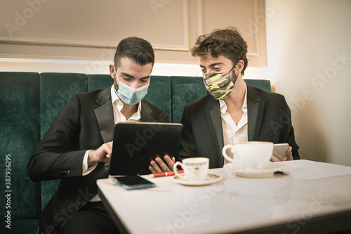 Two young business guys watching contents on a tablet during the breakfast in a hotel hall. They   re wearing a protective mask against the coronavirus  Covid-19  pandemic.