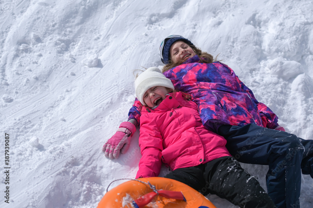 two delightful laughing kids girls wearing ski suit lying on white snow with rubber tube during winter season travel vacation leisure activity with copy space - obrazy, fototapety, plakaty 