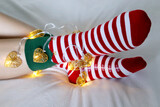 Female legs in Christmas socks with garland and festive lights in heart shape. Concept of romantic New Year night, elf costume