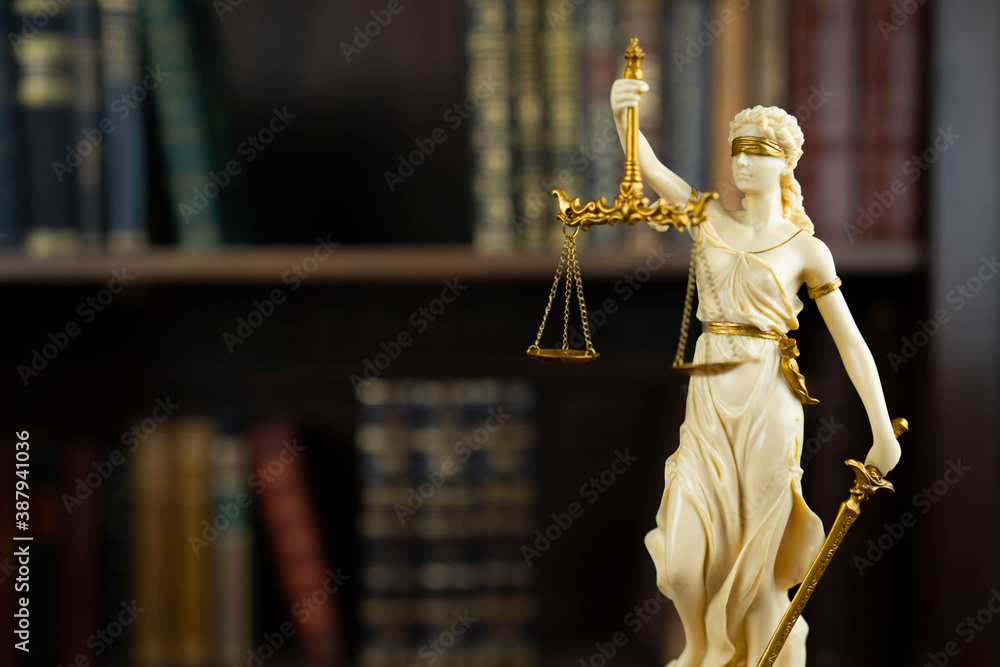 Obraz premium Law and justice concept. Judge chamber. Themis sculpture on brown desk. Collection of legal books in the bookshelf.