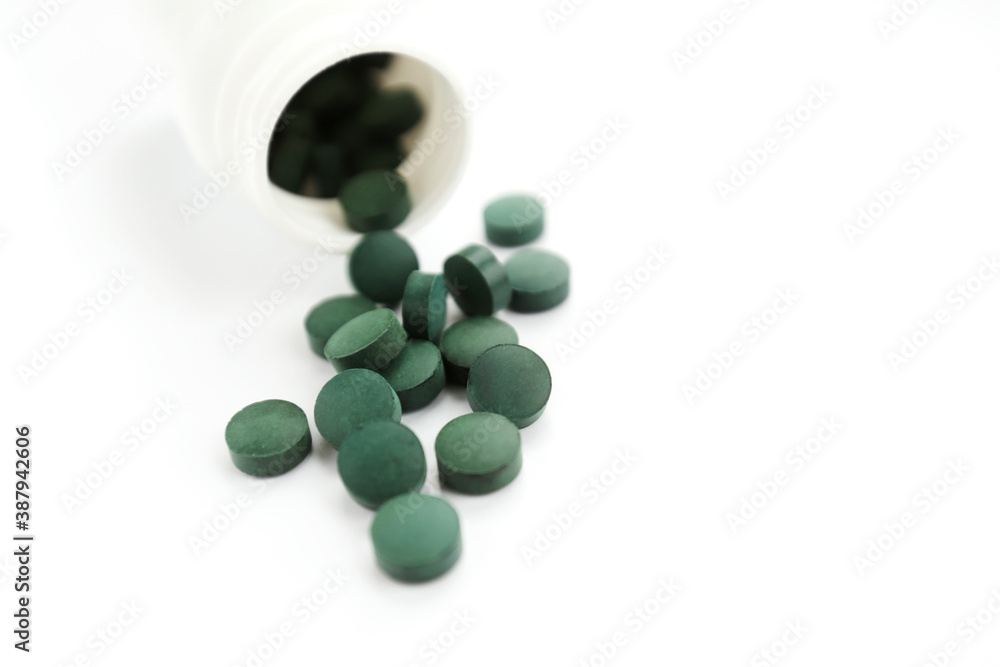 Green capsules of spirulina as healthy food supplements falling from white bottle close-up top view on the white background. Natural medicine and healthcare. Simple template, place for text
