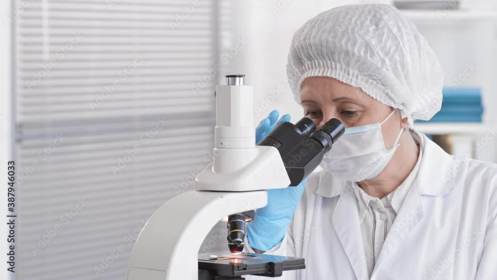 Close-up POV of senior female Asian assistant wearing medical protection working in laboratory, using microscope, taking face mask off looking up on camera, unrecognizable colleagues passing by Close-up POV of senior female Asian assistant wearing medical protection working in laboratory, using microscope, taking face mask off looking up on camera, unrecognizable colleagues passing by