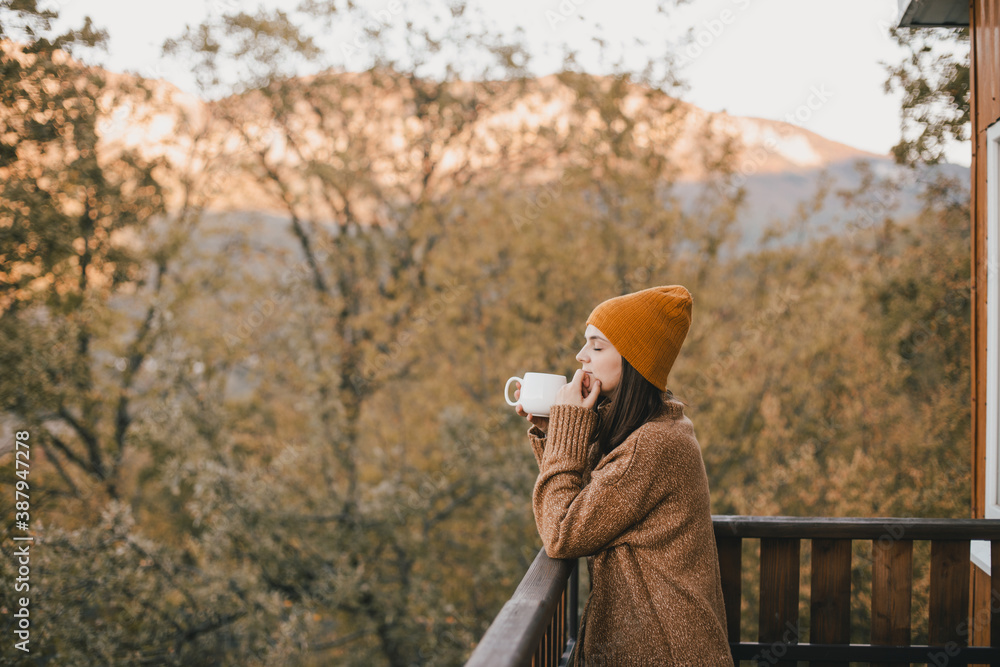 Young woman in knitted sweater and hat drinking tea and eating fresh croissants on cozy balcony of a wooden country house.