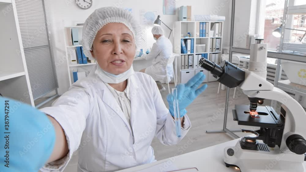 POV of elderly Asian female doctor filming vlog in laboratory, holding test tube with liquid, talking on camera, turning around asking colleague working behind to send greetings to subscribers POV of elderly Asian female doctor filming vlog in laboratory, holding test tube with liquid, talking on camera, turning around asking colleague working behind to send greetings to subscribers