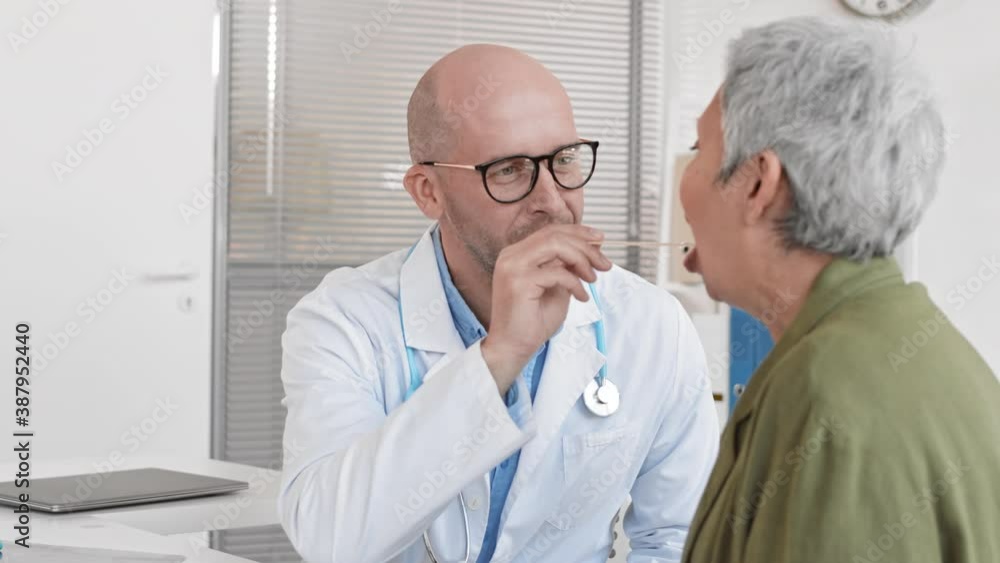 Over-the-shoulder slow motion of middle-aged Caucasian male doctor telling unrecognizable senior patient to open throat widely for checking throat with wooden tongue depressor in clinic office Over-the-shoulder slow motion of middle-aged Caucasian male doctor telling unrecognizable senior patient to open throat widely for checking throat with wooden tongue depressor in clinic office