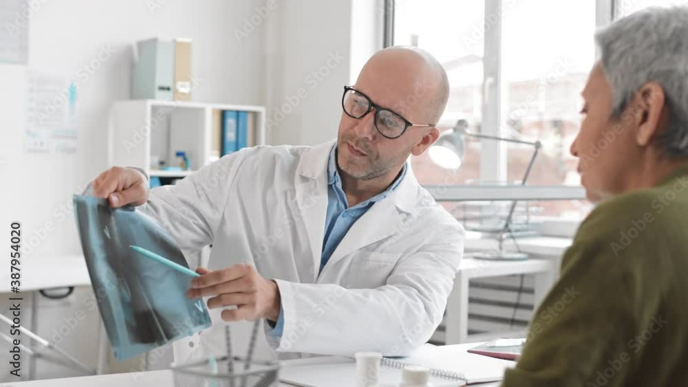 Over-the-shoulder slow motion of middle-aged Caucasian male doctor showing X-rays and talking to cropped senior patient sitting across desk at medical session Over-the-shoulder slow motion of middle-aged Caucasian male doctor showing X-rays and talking to cropped senior patient sitting across desk at medical session