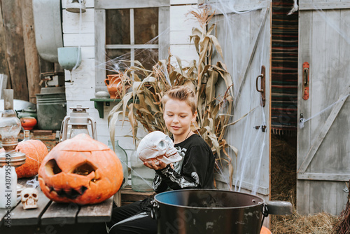 a boy in a skeleton costume with a skull in his hands on the porch of a house decorated to celebrate a Halloween party