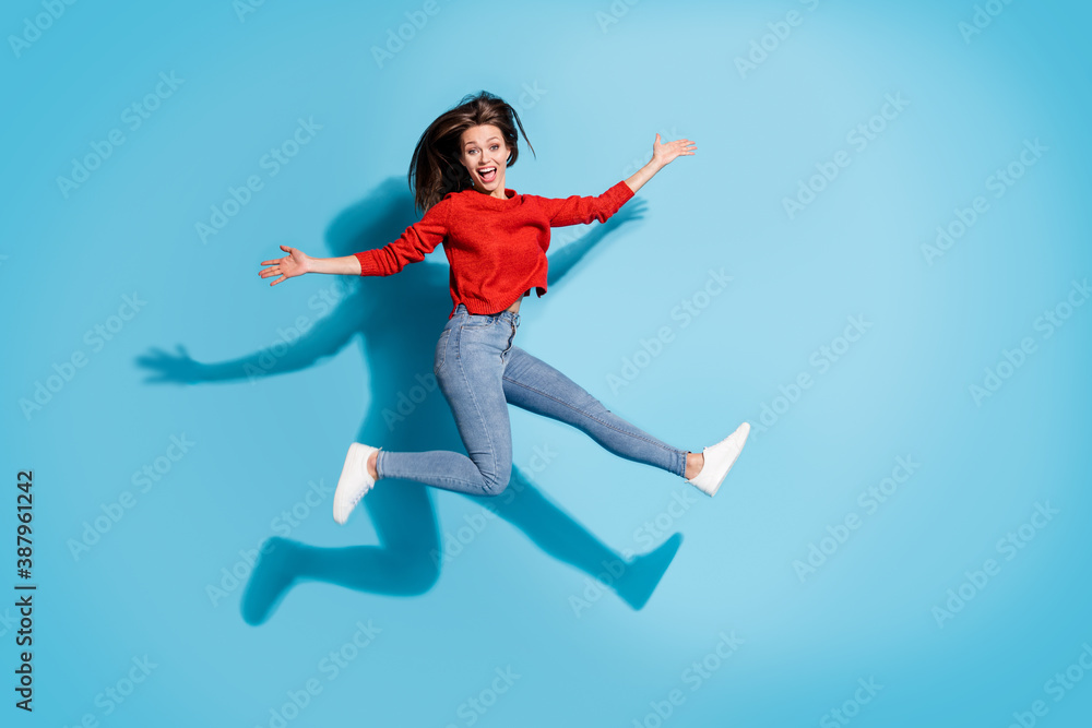 Full length photo of crazy lady jump high up wear knitted red sweater sneakers jeans isolated blue color background Full length photo of crazy lady jump high up wear knitted red sweater sneakers jeans isolated blue color background
