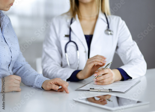 Unknown woman- doctor is listening to her patient, while sitting together at the desk in the cabinet in a clinic. Female physician with a stethoscope is writing at clipboard, close up. Perfect medical