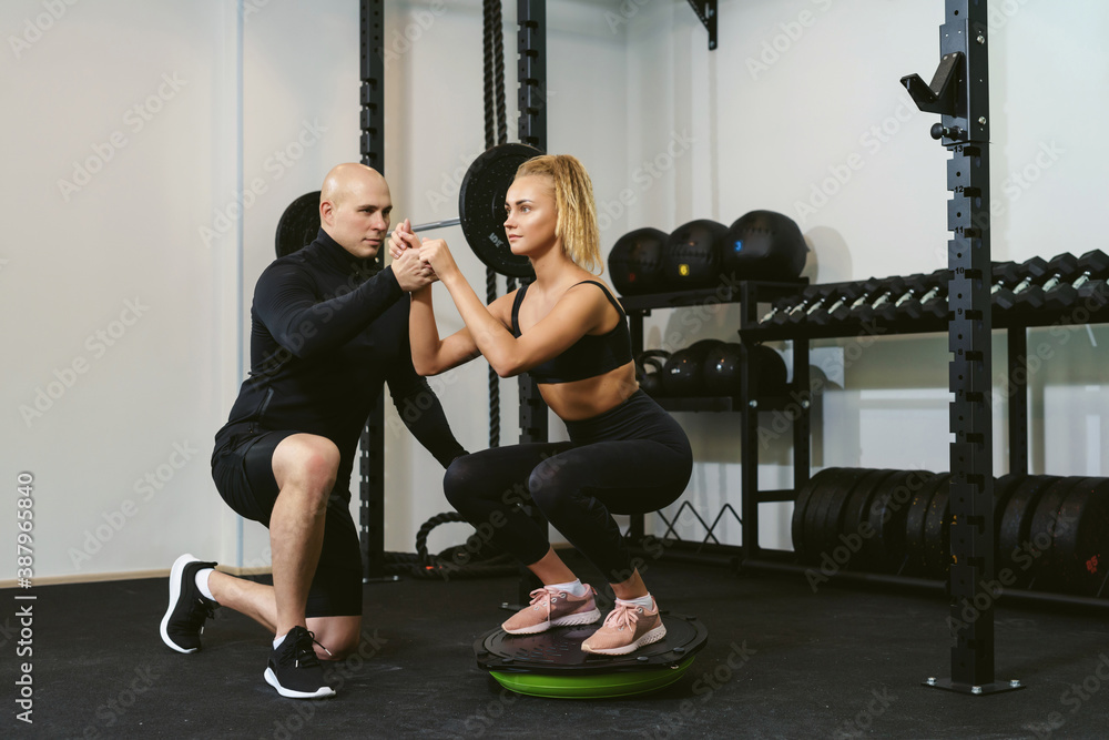 Fototapeta premium A young sportive woman trains butt and legs on a balance board performing lunges and squats. Handsome trainer assisting her and monitoring her sport technique. Personal training with a coach
