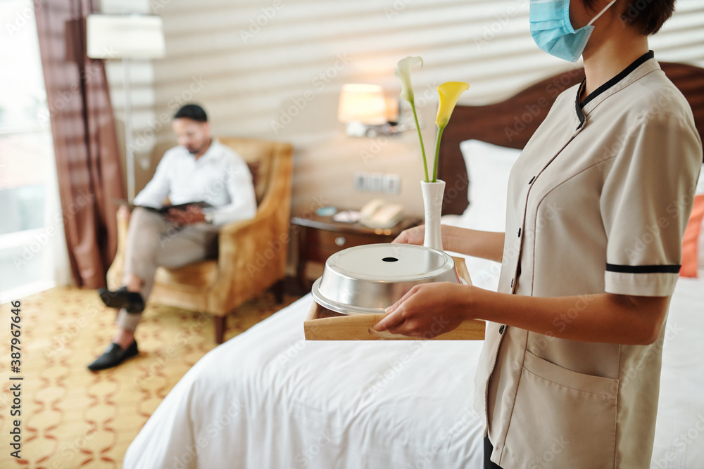 Obraz premium Maid in protective mask bringing hot breakfast and fresh flowers in hotel room of guest