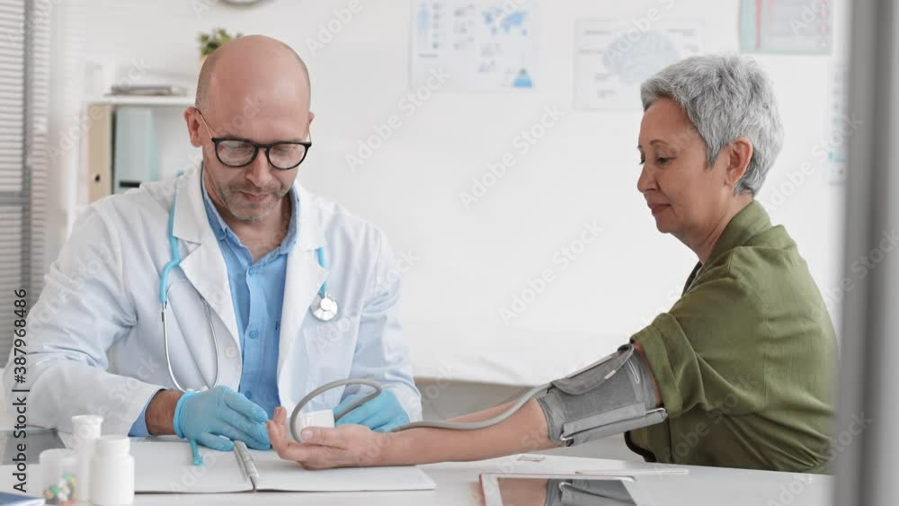Medium slow motion of middle-aged Caucasian male medical worker sitting next to and talking to Asian senior female patient, checking blood pressure with monitor in office Medium slow motion of middle-aged Caucasian male medical worker sitting next to and talking to Asian senior female patient, checking blood pressure with monitor in office