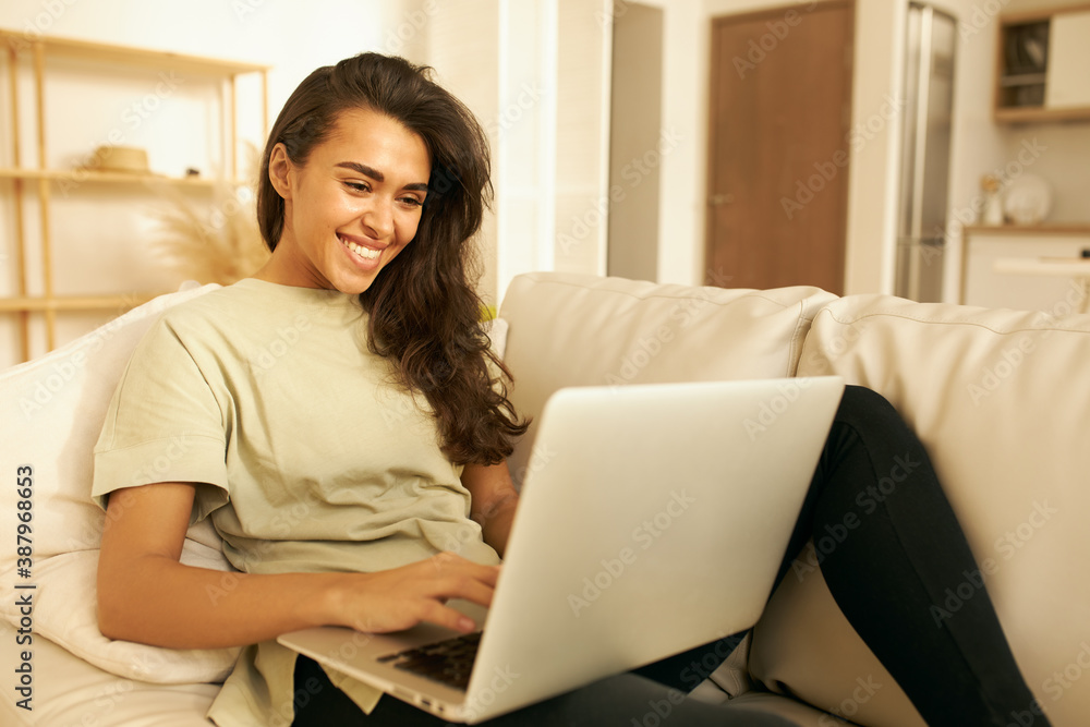 Fototapeta premium Adorable Arabic girl in casual clothes relaxing on couch using laptop, looking at screen with smile, chatting with friends distantly, via group video conference call, enjoying online communication