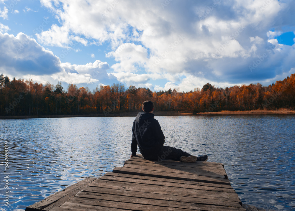 A young man sits on the edge of a wooden bridge near a karst round lake in autumn