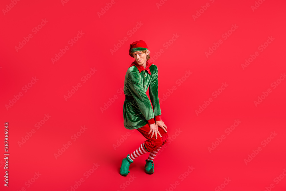 Full size photo of funny elf wearing green costume headwear isolated over red shine color background Full size photo of funny elf wearing green costume headwear isolated over red shine color background