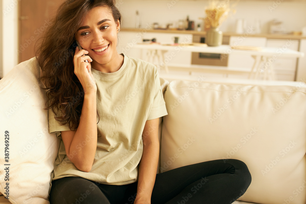 Pretty Latin girl in t-shirt sitting on comfortable sofa, smiling excitedly, receiving good positive news while speaking on phone to friend. Cheerful young female enjoying nice conversation on mobile Pretty Latin girl in t-shirt sitting on comfortable sofa, smiling excitedly, receiving good positive news while speaking on phone to friend. Cheerful young female enjoying nice conversation on mobile