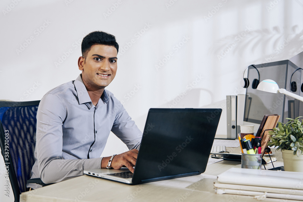 Portrait of smiling Indian programmer working on laptop at office desk Portrait of smiling Indian programmer working on laptop at office desk