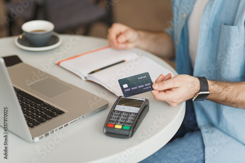 Cropped image of man sit at table in coffee shop cafe restaurant indoor working studying on laptop computer pay off with credit card bank payment terminal. Freelance mobile office business concept.