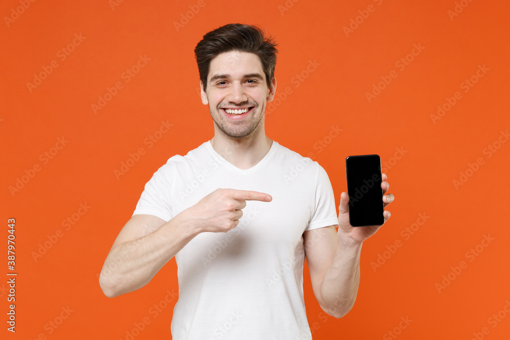 Smiling handsome young man in basic casual white t-shirt pointing index finger on mobile cell phone with blank empty screen looking camera isolated on bright orange colour background studio portrait. Smiling handsome young man in basic casual white t-shirt pointing index finger on mobile cell phone with blank empty screen looking camera isolated on bright orange colour background studio portrait.