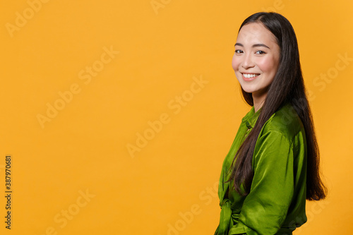 Side view of smiling charming pretty attractive beautiful young brunette asian woman 20s wearing basic green shirt standing looking camera isolated on bright yellow colour background, studio portrait.