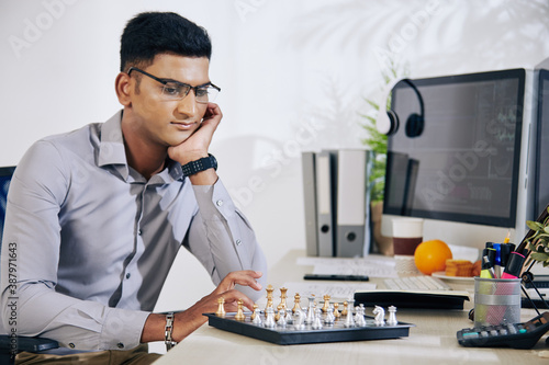 Pensive handsome young Indian software developer looking at chess board and thinking on next move