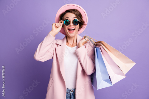 Portrait of amazed adorable sweet brunette hair young lady hold bags touch glasses wear pink cap coat isolated on purple color background