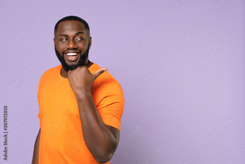 Smiling funny young african american man 20s wearing basic casual orange t-shirt standing pointing thumb aside on mock up copy space isolated on pastel violet colour background studio portrait. Smiling funny young african american man 20s wearing basic casual orange t-shirt standing pointing thumb aside on mock up copy space isolated on pastel violet colour background studio portrait.