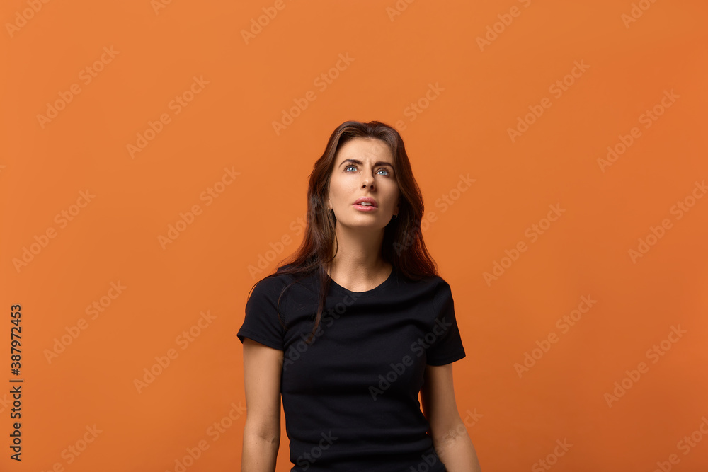 Do you believe in this. Surprised caucasian woman in black t-shirt looks thoughtfully upwards, trying to understand what's there. Isolated over an orange background. Do you believe in this. Surprised caucasian woman in black t-shirt looks thoughtfully upwards, trying to understand what's there. Isolated over an orange background.