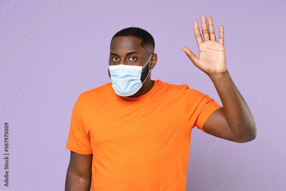 Young african american man 20s wearing casual orange t-shirt sterile face mask to safe from coronavirus virus covid-19 waving greeting with hand isolated on pastel violet background, studio portrait. Young african american man 20s wearing casual orange t-shirt sterile face mask to safe from coronavirus virus covid-19 waving greeting with hand isolated on pastel violet background, studio portrait.