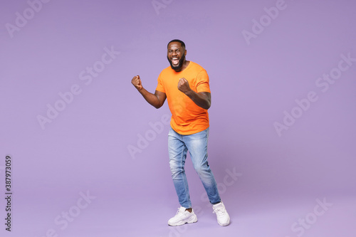 Full length of screaming young african american man 20s wearing basic casual orange t-shirt clenching fists doing winner gesture looking camera isolated on pastel violet background studio portrait.