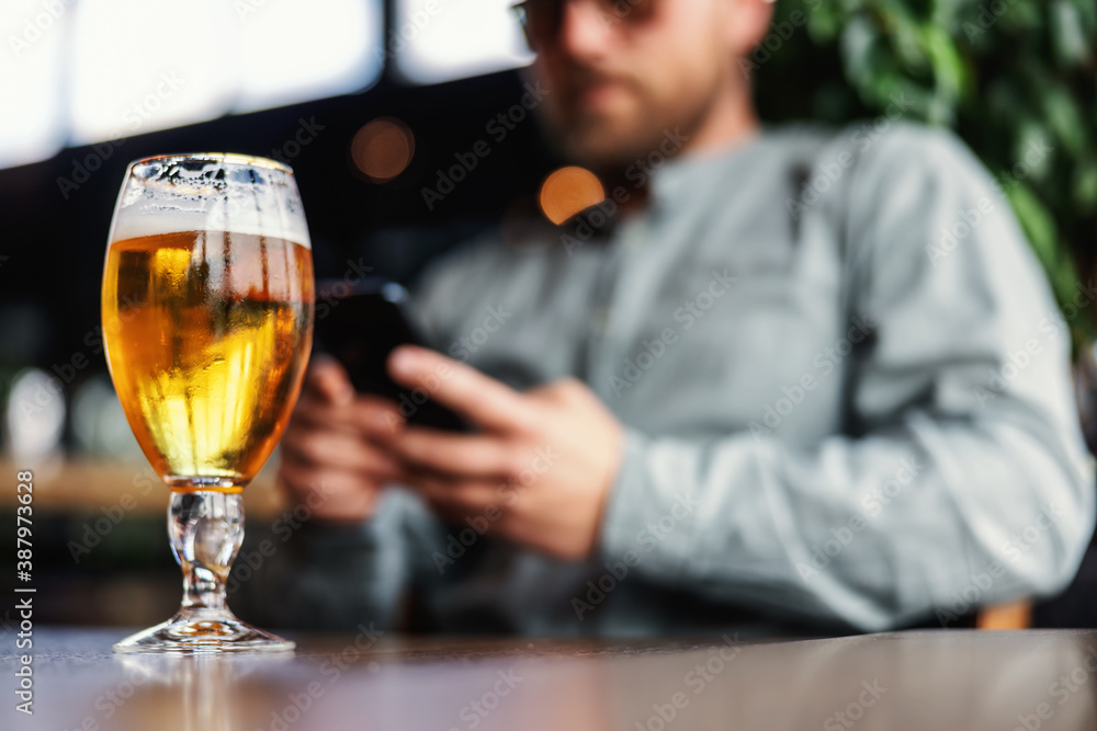 Closeup of fresh cold light beer in glass. In background is man texting message on the phone.