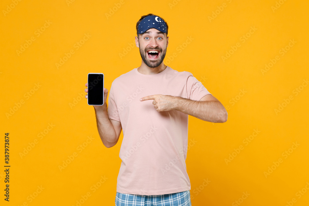 Surprised young man in pajamas home wear sleep mask pointing index finger on mobile cell phone with blank empty screen resting at home isolated on yellow background. Relax good mood lifestyle concept. Surprised young man in pajamas home wear sleep mask pointing index finger on mobile cell phone with blank empty screen resting at home isolated on yellow background. Relax good mood lifestyle concept.