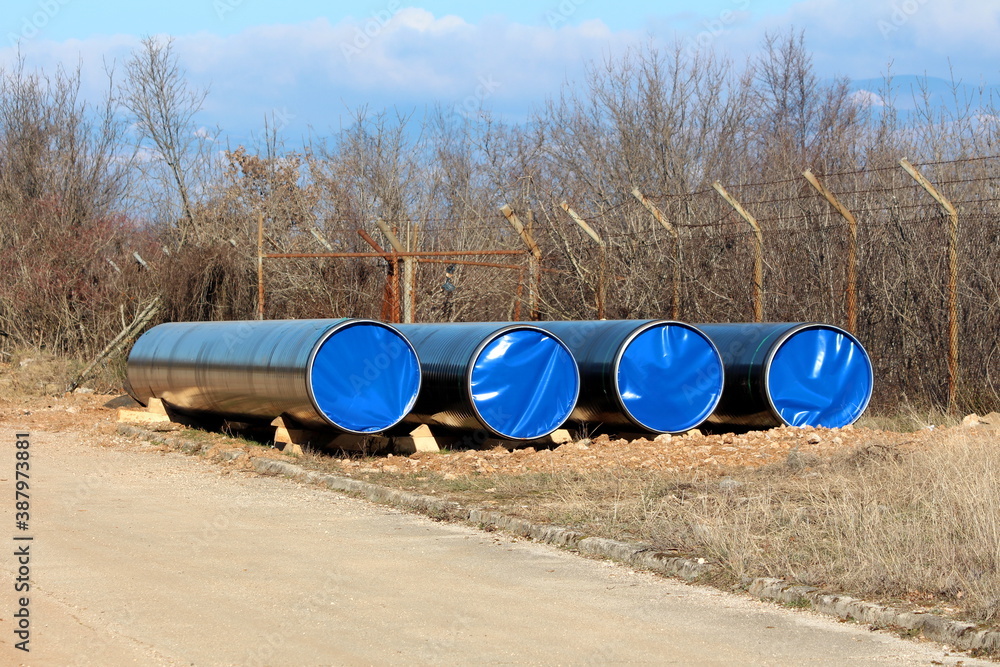 Four brand new large diameter pipes closed with nylon protection left at local oil pipeline construction site next to paved road and wire fence with barb wire on top surrounded with dense trees withou Four brand new large diameter pipes closed with nylon protection left at local oil pipeline construction site next to paved road and wire fence with barb wire on top surrounded with dense trees withou