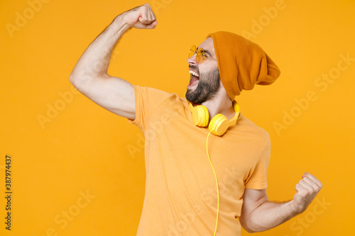 Overjoyed screaming young bearded man 20s wearing basic casual t-shirt headphones eyeglasses hat standing clenching fists doing winner gesture isolated on bright yellow background, studio portrait.