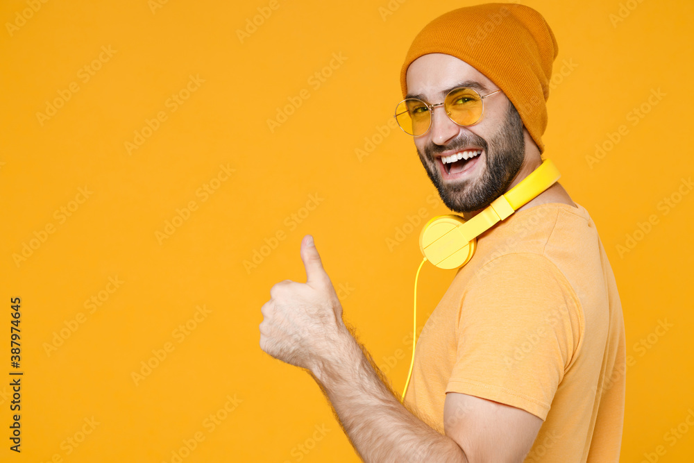 Fototapeta premium Side view of cheerful funny young bearded man 20s wearing basic casual t-shirt headphones eyeglasses hat standing showing thumb up looking camera isolated on bright yellow background, studio portrait.