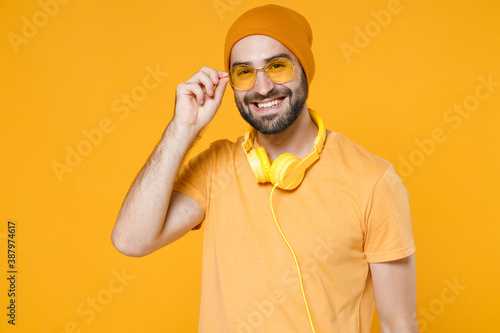 Smiling cheerful handsome attractive young bearded man 20s wearing basic casual t-shirt headphones eyeglasses hat standing looking camera isolated on bright yellow colour background, studio portrait.