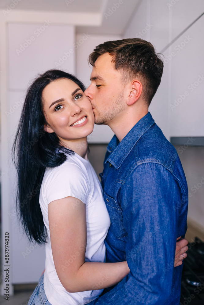 A young man kisses his girlfriend, hugging while standing in the kitchen. The couple hugs.